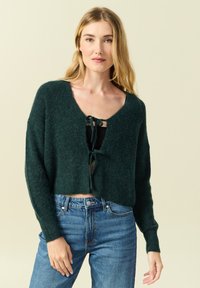 Cardigan verde a maglia corto con scollo a V e chiusura frontale legata, dotato di maniche lunghe e di un tessuto morbido e texturizzato, abbinato a jeans blu.