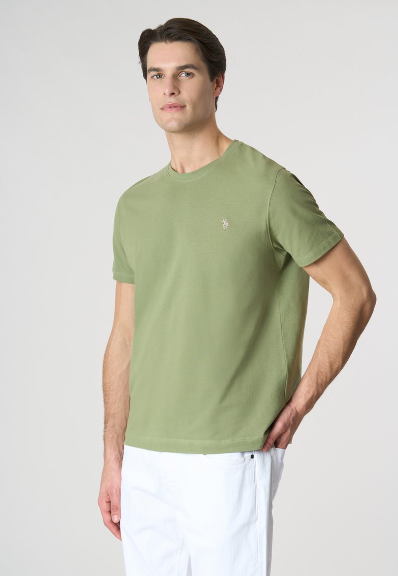 T-shirt in cotone verde oliva a maniche corte con scollatura rotonda e un piccolo logo ricamato sul petto sinistro. Abbinato a pantaloni bianchi.