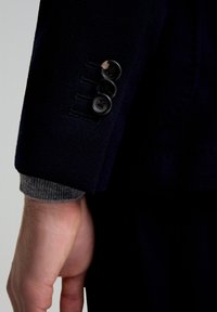 Manga de chaqueta de traje texturizado en azul marino con tres botones negros y puño de punto gris. Los detalles incluyen costuras y un corte entallado.