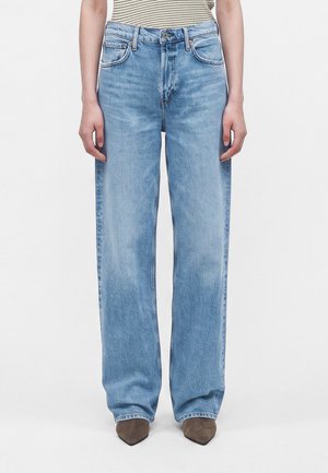 Person iført lyseblå vidde jeans og brune spidse sko, der står mod en ensfarvet hvid baggrund.
