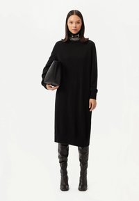 Svart stickad klänning med hög halsringning, långa ärmar och ribbade muddar, kombinerad med en texturerad svart clutch och knähöga svarta stövlar.