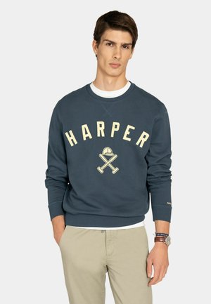 Joven con sudadera azul marino con el texto "HARPER" y un diseño de bates y gorra de béisbol cruzados, pantalones beige y un reloj de pulsera, con las manos en los bolsillos.