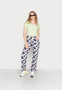 Camisa verde de manga curta com botões; calças largas estampadas com quadrados em roxo, preto e branco; ténis brancos com detalhes amarelos.