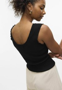 Top sin mangas negro con textura acanalada y bordes en forma de onda, con un ajuste entallado y un escote pronunciado en la espalda, combinado con pantalones claros de textura acanalada.
