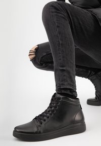 Černé vysoké tenisky vyrobené z hladké kůže, s kulatou špičkou, plochými tkaničkami a texturovanou gumovou podešví, spárované s roztrhanými černými džíny.