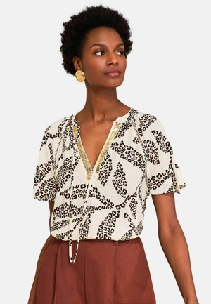 Femme portant un chemisier à imprimé léopard avec bordure dorée et un pantalon marron taille haute, regardant sur le côté avec des boucles d'oreilles circulaires dorées.