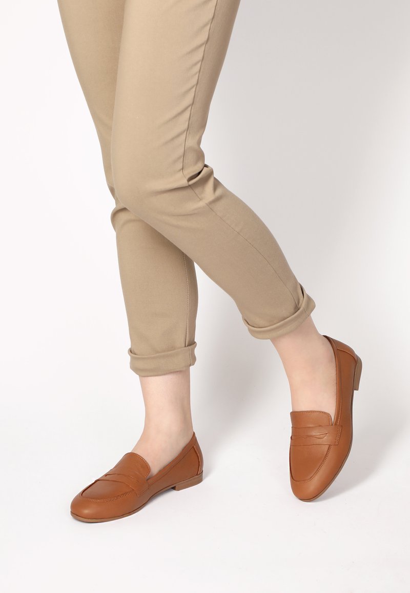 Braune Lederloafers mit einem gestickten Design, niedrigem Absatz und glatter Textur. Kombiniert mit hochgekrempelten beigen Hosen.