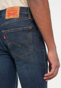 Gros plan d'une personne portant un jean Levi's 511 bleu foncé avec une poche arrière visible, une étiquette de marque et le bras droit partiellement étendu.