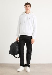 Felpa bianca con cappuccio, coulisse e tasca frontale, abbinata a jeans neri. Modello tiene una borsa nera, indossando sneaker bianche. Look semplice e casual.
