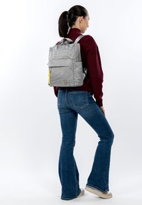 Grijze rugzak met een gestructureerd ontwerp, meerdere compartimenten en een gele ritssluiting, gedragen op de rug over een wijnrode trui en blauwe spijkerbroek.