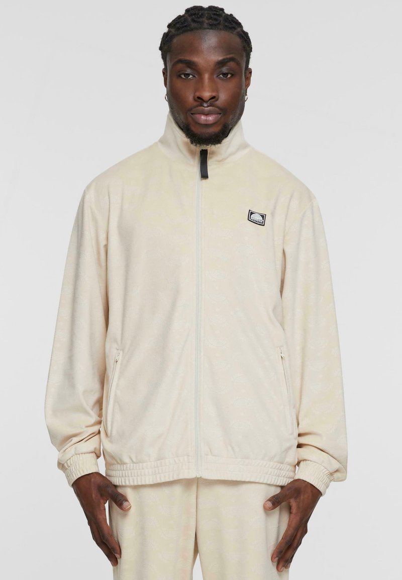 Veste zippée beige clair avec col haut, poignets côtelés et poches latérales, arborant un motif subtil et un petit logo sur la poitrine.