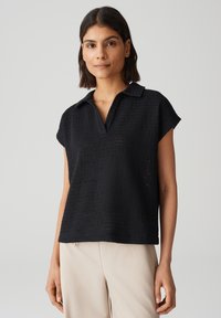 Polo corto in maglia nera a maniche corte con scollo a V, dotato di un motivo texture e colletto classico. Abbinato a pantaloni di colore chiaro.