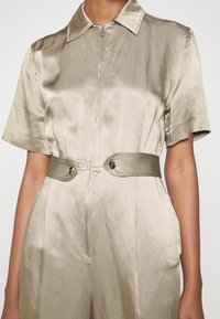 Combinaison à manches courtes en satin vert olive, avec un zip à l'avant, un large col et une taille ceinturée avec des accents métalliques. Texture lisse.