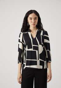 Blusa nera e crema con un motivo geometrico, scollatura a V, maniche a tre quarti e dettagli arricciati ai polsini. Indossata sopra pantaloni neri.
