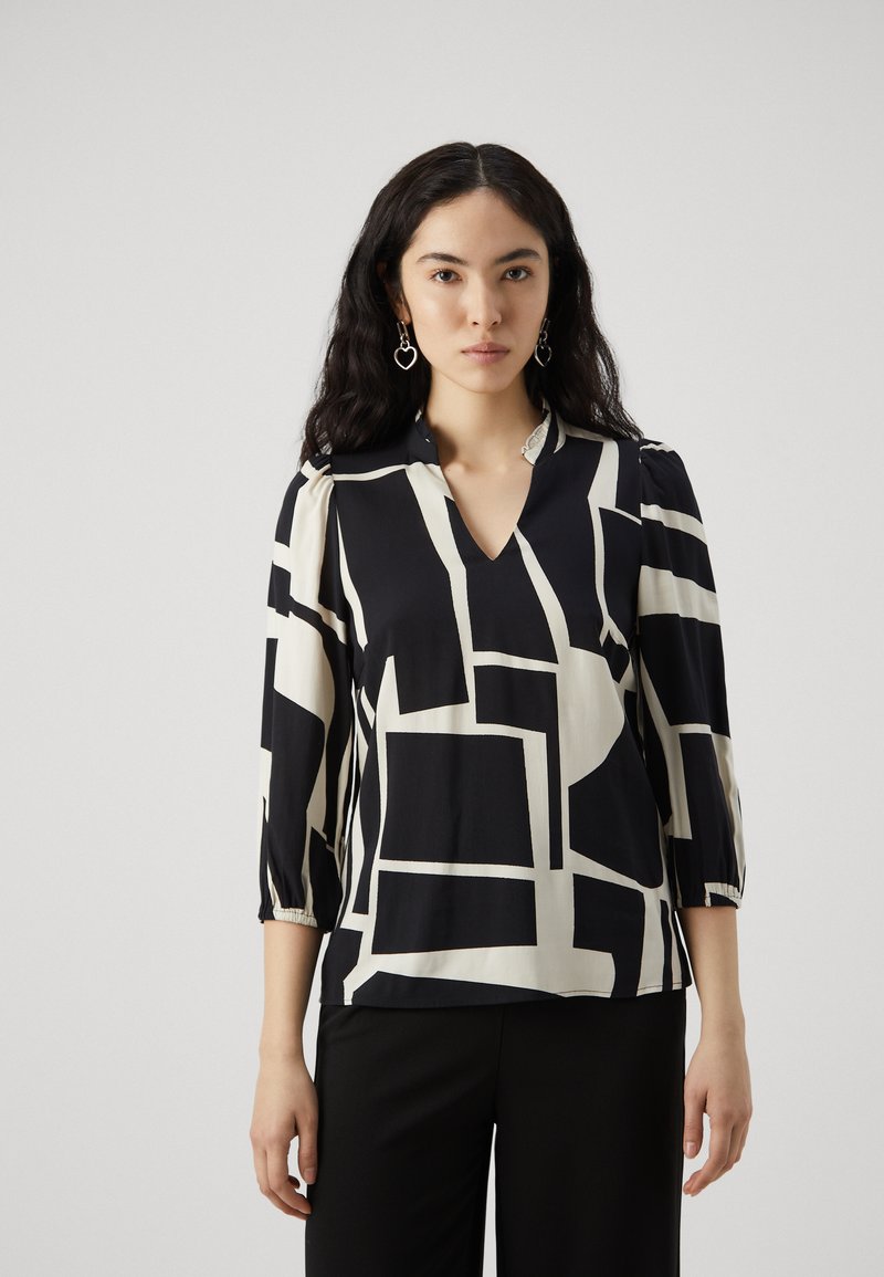 Blusa nera e crema con un motivo geometrico, scollatura a V, maniche a tre quarti e dettagli arricciati ai polsini. Indossata sopra pantaloni neri.