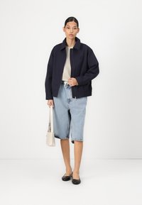 Jachetă de bombardier navy cu închidere cu fermoar, top bej, pantaloni scurți din denim bleu deschis și pantofi negri cu talpă joasă. Ținând o poșetă de culoare deschisă.