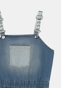 Džínový overal s nastavitelnými popruhy a potiskem "DRESS CODE". Světle modrá barva, kovové spony a detail na zadní kapse. S nařaseným pasem.