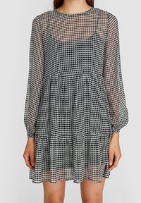 Vestido de houndstooth en blanco y negro con transparencias, de manga larga, escote redondo y falda con volantes, que presenta una textura ligera.