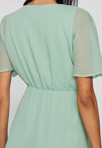 Een vrouw in een lichtgroene, gestructureerde jurk met korte doorzichtige mouwen en een elastische taille, gezien vanuit de rug.