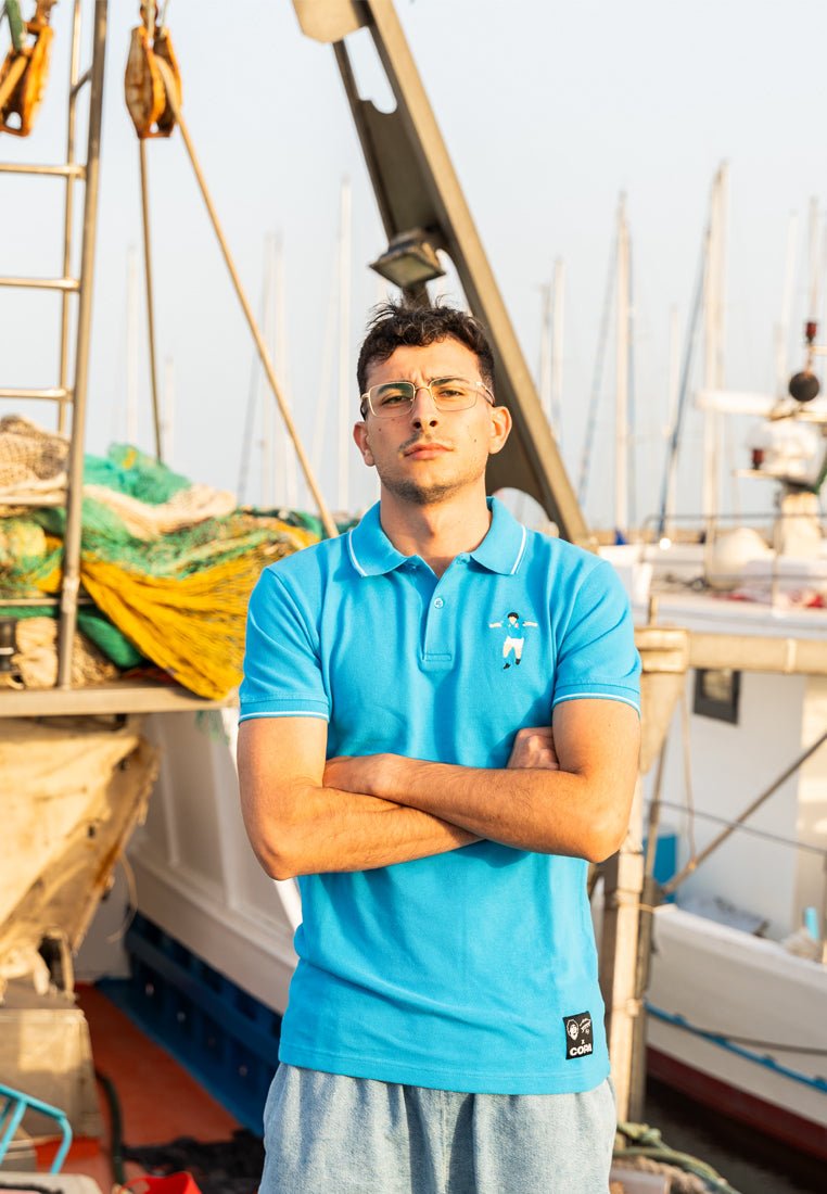 Turquoise poloshirt met korte mouwen en een klein geborduurd ontwerp op de borst, gecombineerd met lichtgrijze shorts. De achtergrond toont boten en vistuig.