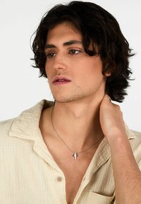 Ciondolo a croce d'argento su una catena sottile, indossato sulla pelle nuda; il soggetto ha i capelli corti e mossi di colore scuro ed è vestito con una camicia beige a trama.