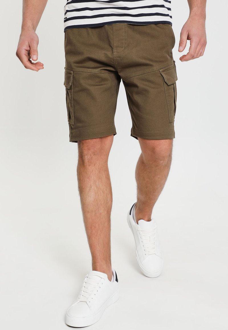 Pantaloni cargo verde oliva con tasche laterali, orlo arrotolato e tessuto di cotone resistente. Abbinati a sneaker bianche. Camicia a righe orizzontali visibile.