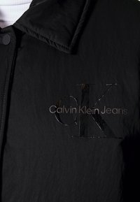 Sort jakke med en tekstureret stof, en spids krave og et delvist glanset logo prægede foran, som læser "Calvin Klein Jeans."