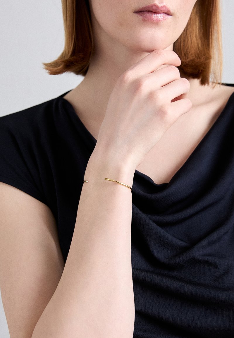 Χρυσό μεταλλικό βραχιόλι με λεπτή ταινία και μικρή στρογγυλή λεπτομέρεια, φορεμένο στον καρπό, σε φόντο σκούρου υφάσματος. Απλός και μινιμαλιστικός σχεδιασμός.