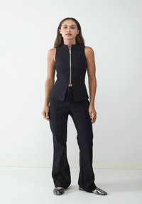 Zwarte mouwloze top met een hoge kraag en ritssluiting aan de voorkant, gecombineerd met zwarte flared broeken en metallic platte schoenen, tegen een eenvoudige achtergrond.