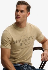 Camiseta beige de manga corta con cuello redondo, con un estampado oscuro que dice "SUPERDRY & CO" y "NEW YORK." Textura suave, ajuste casual.