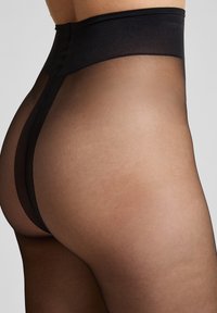 Nahaufnahme einer Person, die durchsichtige schwarze Strumpfhosen mit verstärktem schwarzen Bund und rückseitiger Nahtdetails trägt.