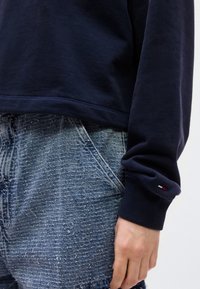 Φούτερ σε σκούρο μπλε ναυτικό χρώμα με ριμπ μανσέτες, συνδυασμένο με ανοιχτό μπλε τζιν υφής που διαθέτει μπροστινή τσέπη. Διακριτικό λογότυπο στο μανίκι.