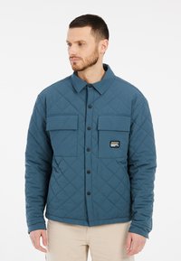 Gequiltede blauwe jas met twee borstzakken, zwarte knopen en een geborduurd logo aan de linkerkant. Gladde textuur, casual ontwerp.