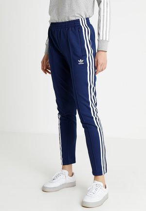 Osoba ubrana w granatowe spodnie dresowe Adidas z białymi paskami, szarą bluzkę z długim rękawem i białe sneakersy, stojąca na jednolitym tle.