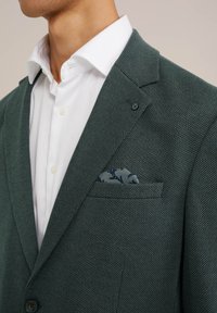 Green textured blazer with a single button, white shirt underneath, and a blue patterned pocket square in the breast pocket.