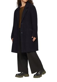 Navyblauer Fischgrätmantel mit Reverskragen, Ein-Knopf-Verschluss und seitlichen Taschen, kombiniert mit einem braunen Oberteil und schwarzen, weiten Hosen.