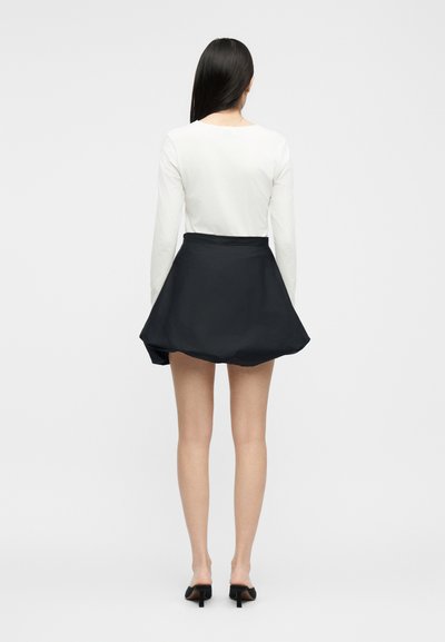 Femme aux longs cheveux foncés portant un haut blanc à manches longues, une mini-jupe noire évasée et des mules à talons noires, debout de dos sur fond blanc.