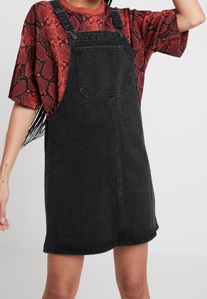 Musta farkku overall-tyylinen mekko, jossa on säädettävät hihnat, etutasku ja suora leikkaus. Yllä punainen käärmeennahkakuosisessa T-paidassa.