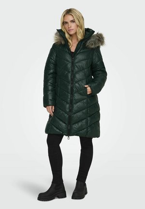 Doudoune matelassée verte avec une capuche en fourrure, dotée d'une fermeture éclair sur le devant et de surpiqûres en biais. Portée avec un pantalon noir et des bottes.