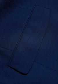 Granatowy materiał o gładkiej fakturze, z płaską kieszenią wkomponowaną w projekt. Widać czyste linie i minimalne detale.