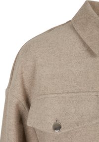 Béžová vlněná bomber bunda s límcem a přední kapsou, která má stříbrný knoflíkový detail. Měkká textura a volný střih.