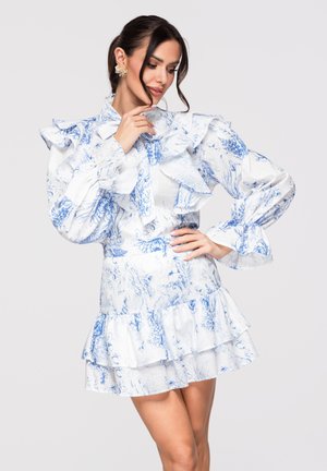 Donna che indossa un vestito bianco e blu a fiori con balze, maniche lunghe a sbuffo e gonna a strati, posando con la mano sul mento e sul fianco.
