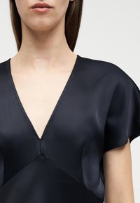 Κοντινό πλάνο γυναίκας που φοράει μαύρο σατινέ V-λαιμόκοψη και κοντομάνικο τοπ, δείχνοντας το λαιμό και το επάνω μέρος του στήθους σε απλό φόντο.