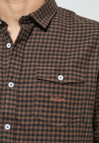 Chemise en flanelle à carreaux marron et noir avec col, présentant une poche poitrine et un logo brodé en orange. Des boutons blancs ornent le devant.