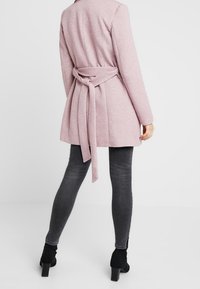 Blush roze jas met een strikceintuur in de taille, gemaakt van textuurstof. Draag het over zwarte skinny jeans en enkellaarsjes. Achteraanzicht zichtbaar.
