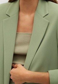 Vaaleanvihreä räätälöity bleiseri lovikauluksilla, jonka alla on beige ribbineulospaita. Sileä kangas, jossa on jäsennelty muoto ja hillitty käden asento bleiserissä.