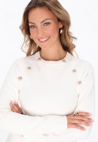 Hvid strikket sweater med rund halsudskæring, guldknap-detaljer, tætsiddende pasform og blød tekstur. Modellen bærer en ring og har lyst, bølget hår.