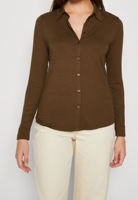 Camisa marrón de manga larga con cuello, abertura frontal con botones y dobladillo redondeado. Hecha de material suave, combinada con pantalones de color crema.