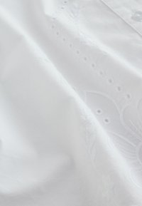 Fehér pamut szövet, bonyolult virágos hímzéssel és texturált részletekkel, gombos záródással és puha, sima felülettel.