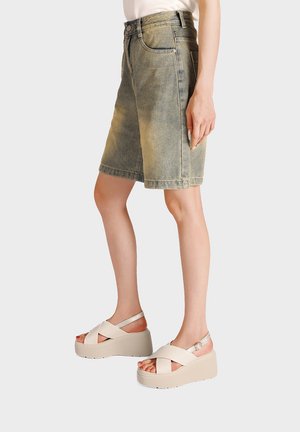 Persona che indossa pantaloncini di jeans sbiaditi fino al ginocchio e sandali beige con plateau e cinturini incrociati, mostrata dalla metà della coscia fino ai piedi.